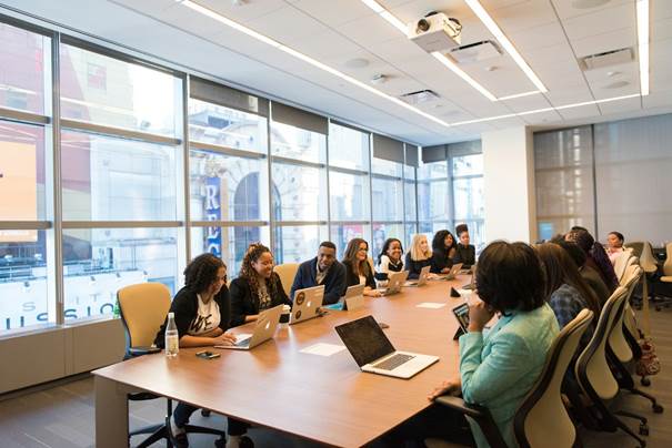 The image depicts a group of people seated around a long table in a modern, well-lit office environment, with large windows providing natural light and a view of the outside.

KI-generierte Inhalte können fehlerhaft sein.