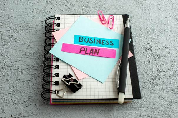 The image depicts a spiral notebook with a pen, a rubber band, and a paper sheet, all decorated with colorful sticky notes, symbolizing a creative and organized business planning session.

KI-generierte Inhalte können fehlerhaft sein.